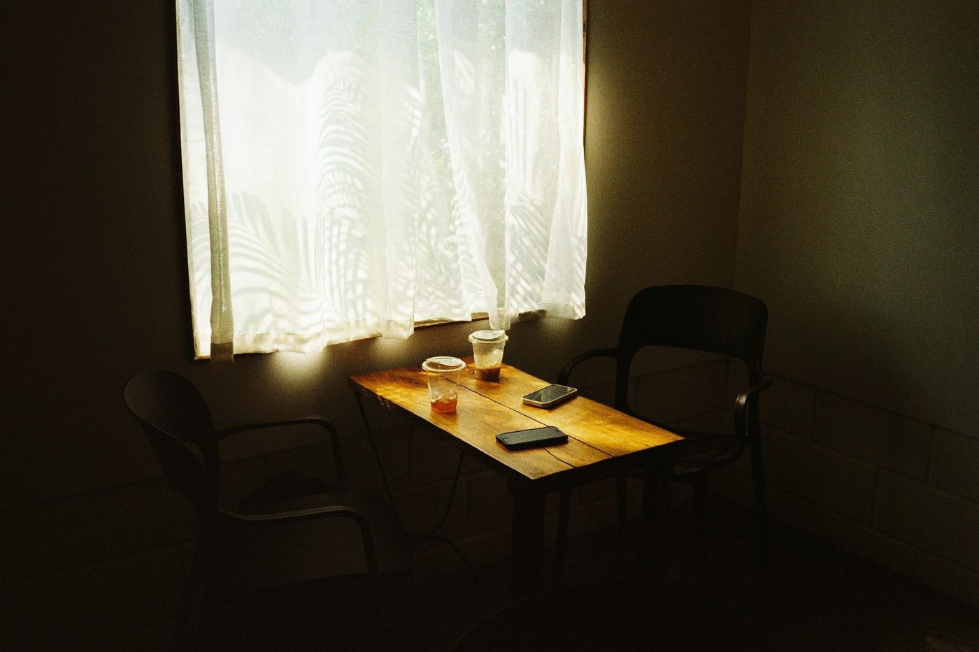 Two empty chairs at a small table by a curtained window, two drinks and two phones face down