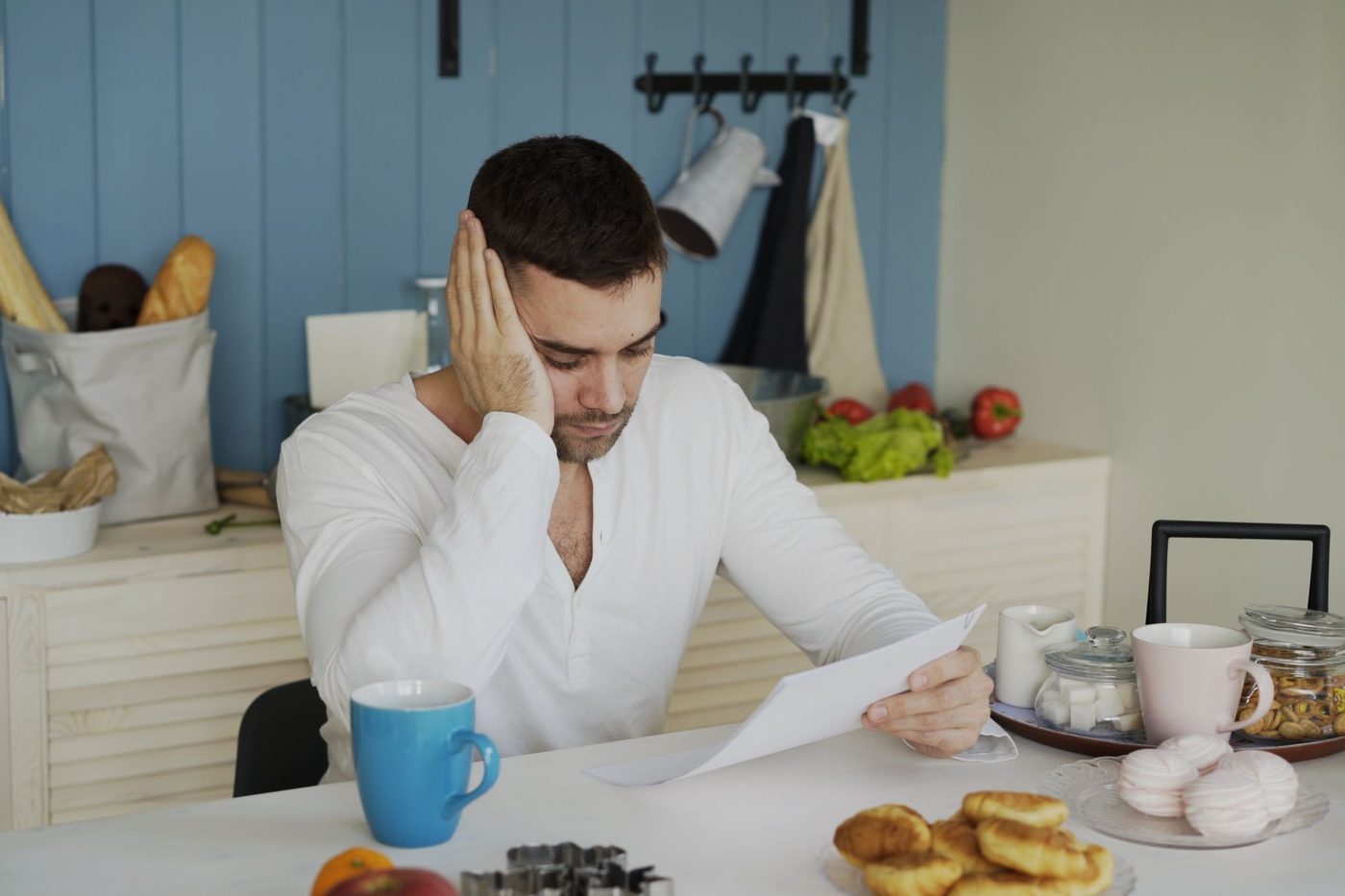 A man at a kitchen table with his hand to his head, reading a printed page