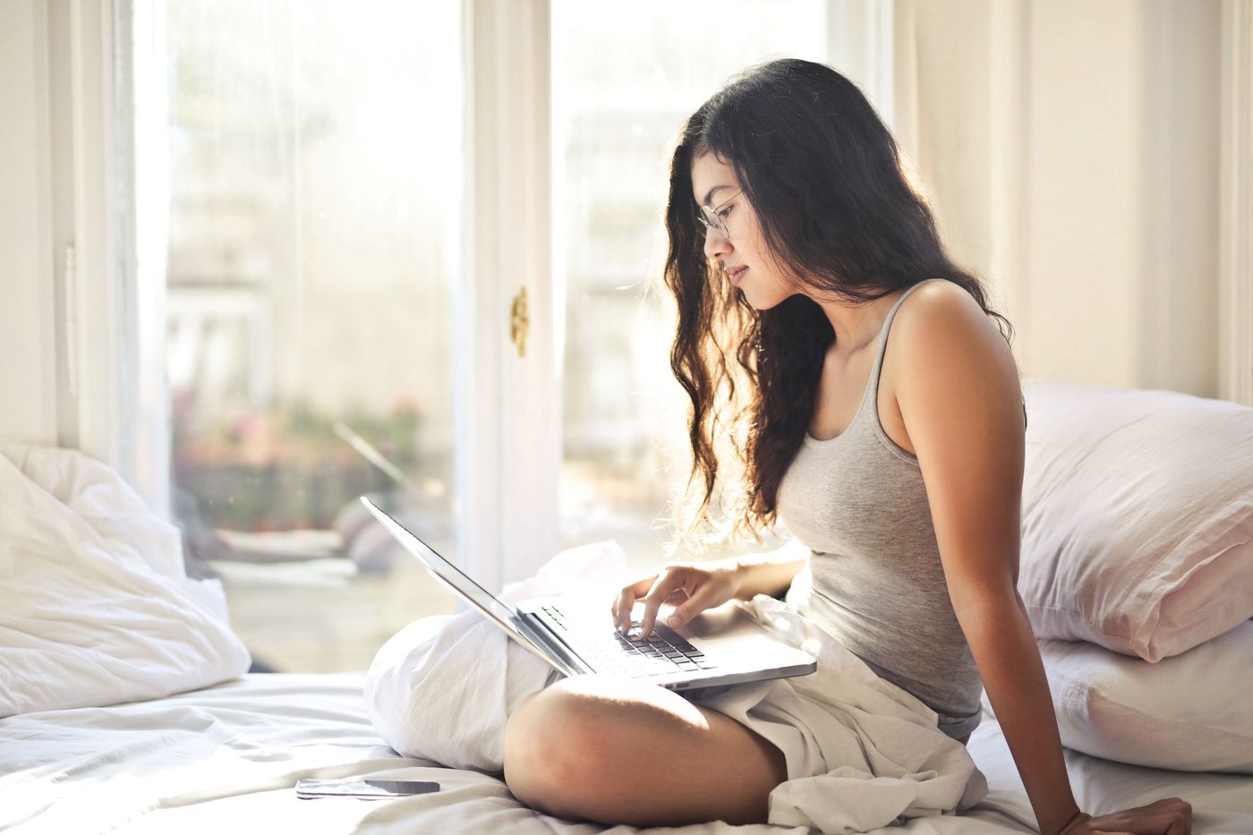 A woman with long dark hair sits cross-legged on a bed in a sunlit room, typing on a laptop