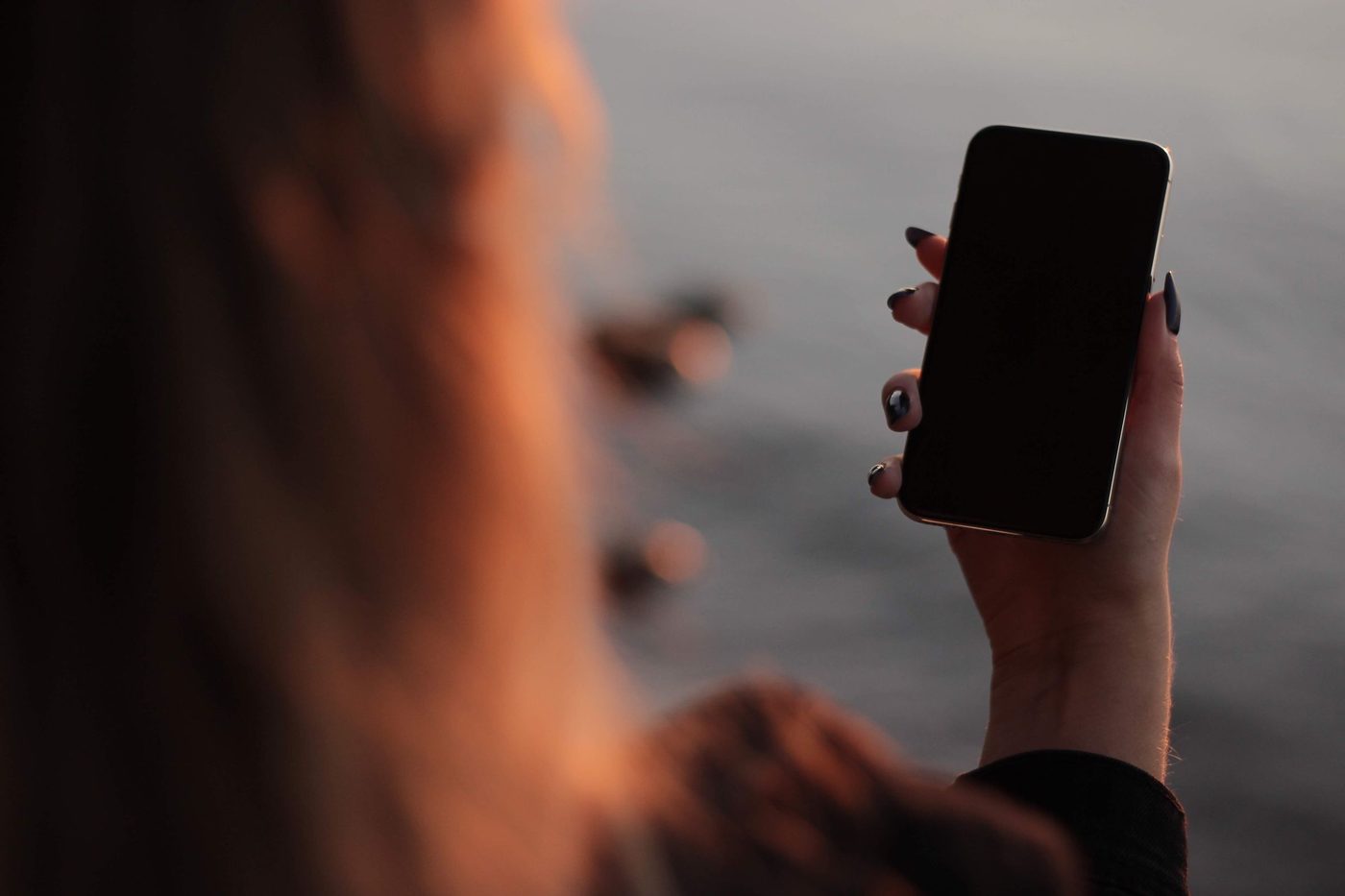 A hand holding a phone with a dark screen, a woman looking out over water at dusk