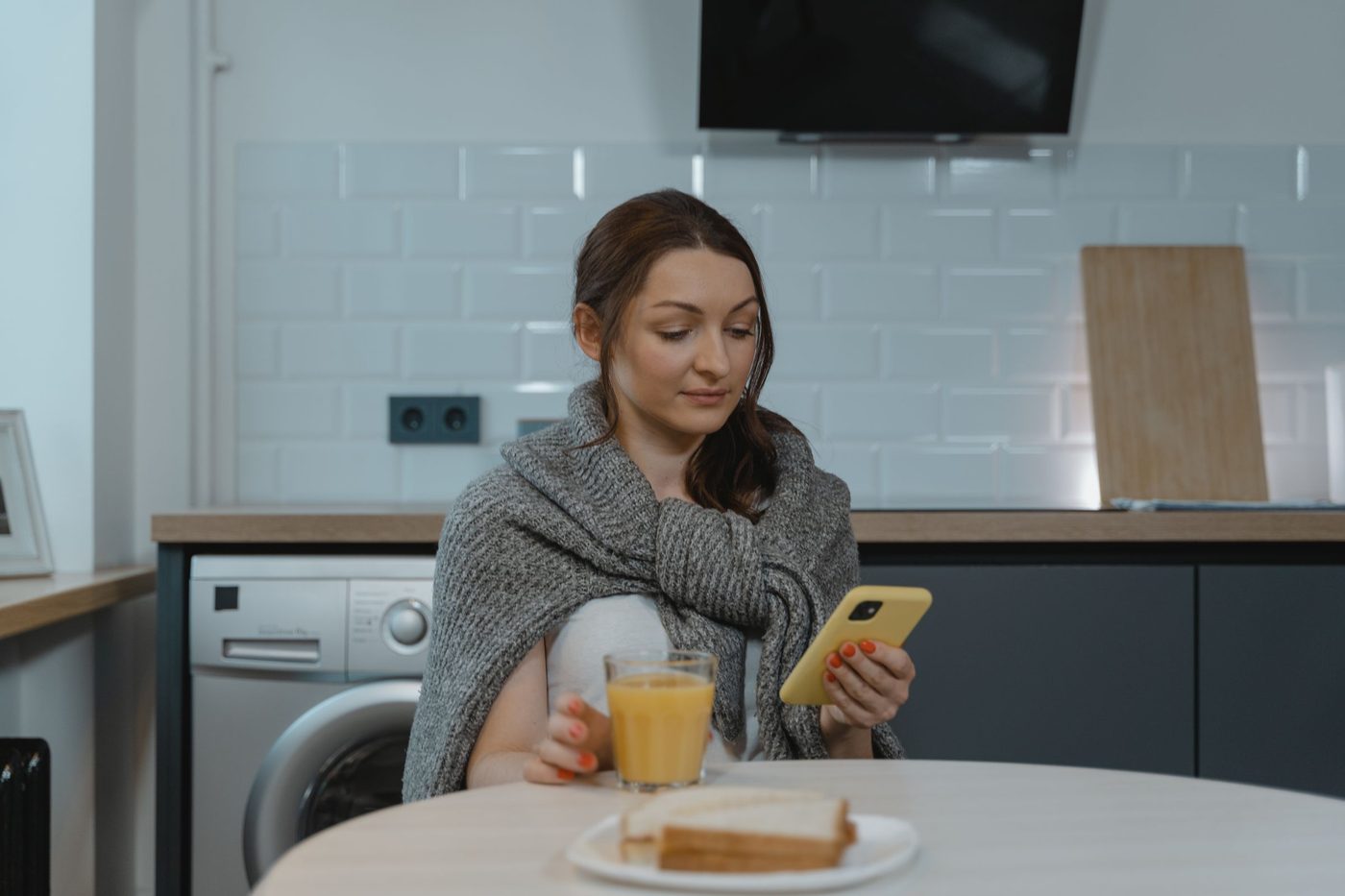 A woman in a grey knitted cardigan looks at her phone at a kitchen table