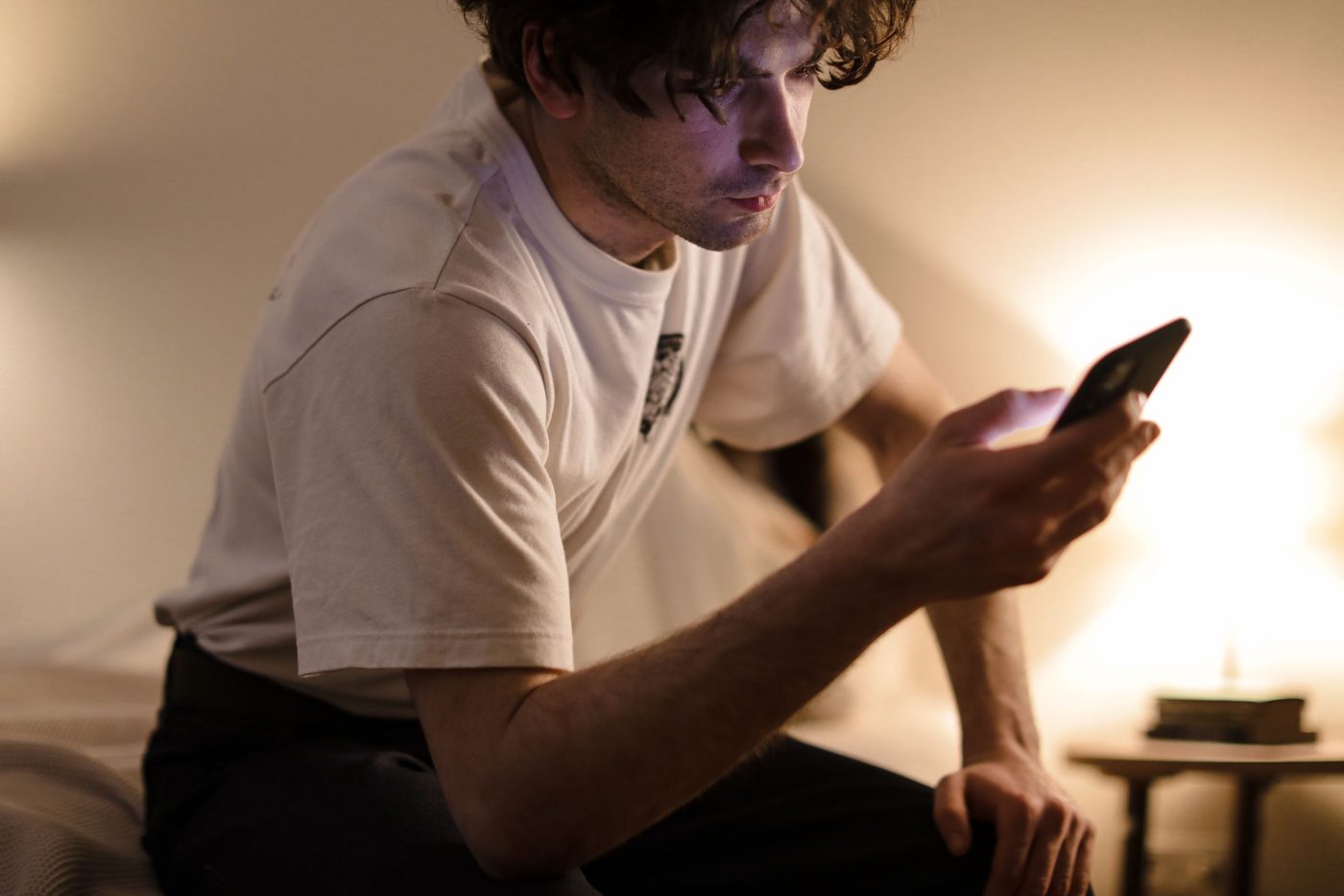 A man sits on the edge of a bed in warm lamp light, reading his phone
