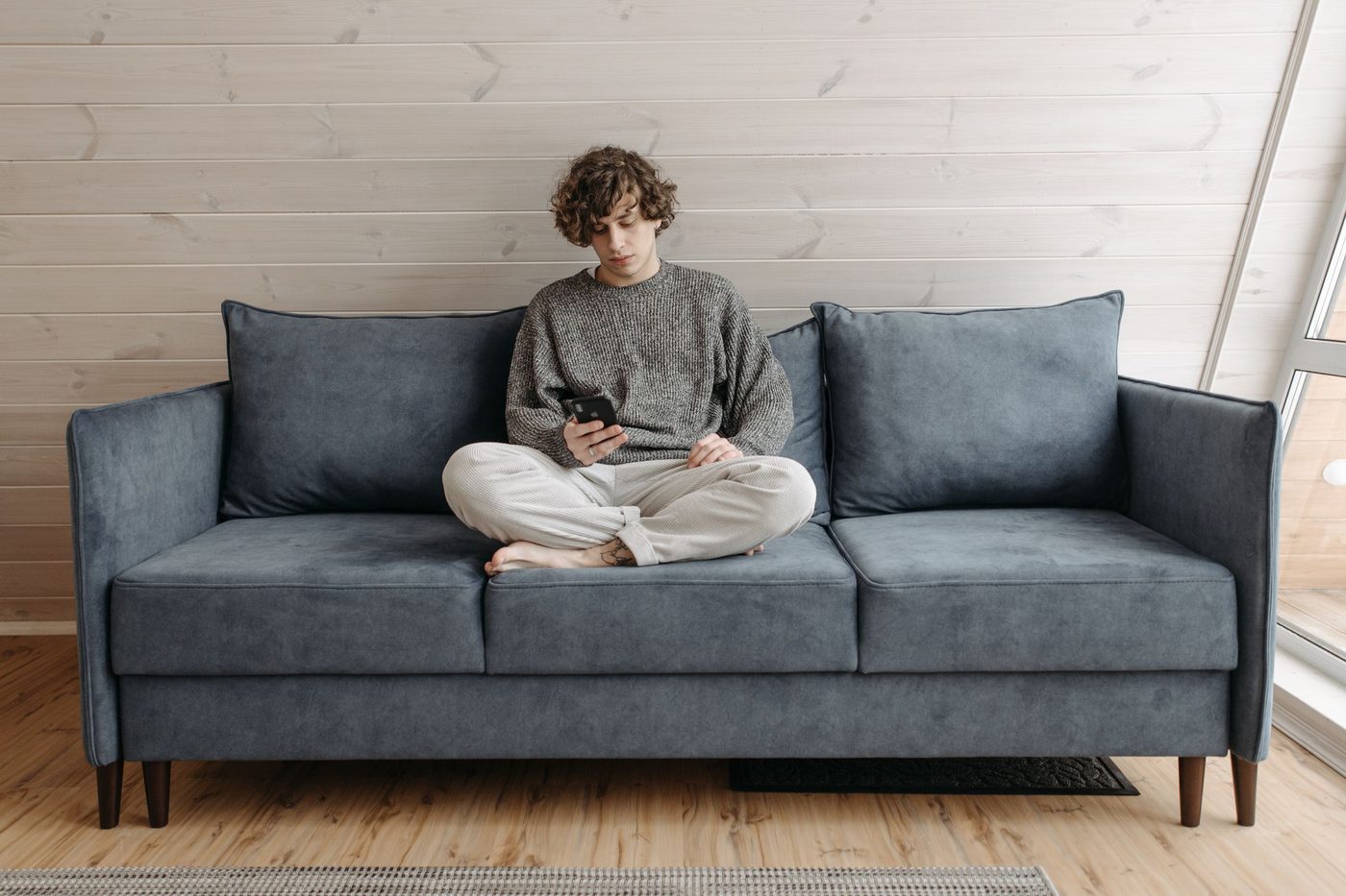 A young man sits cross-legged on a blue sofa in a bright room, looking at his phone