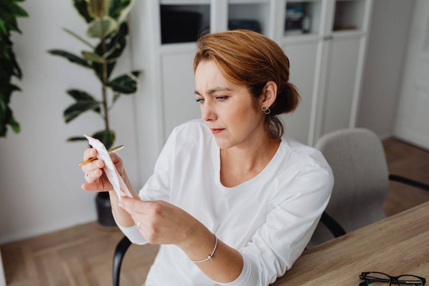 A woman in a white top frowns as she reads a receipt at a wooden desk