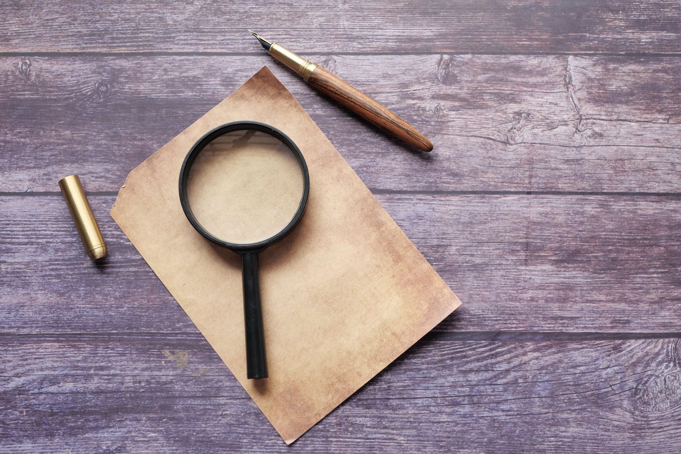 A black-framed magnifying glass rests on a sheet of aged paper beside a fountain pen on a wooden table