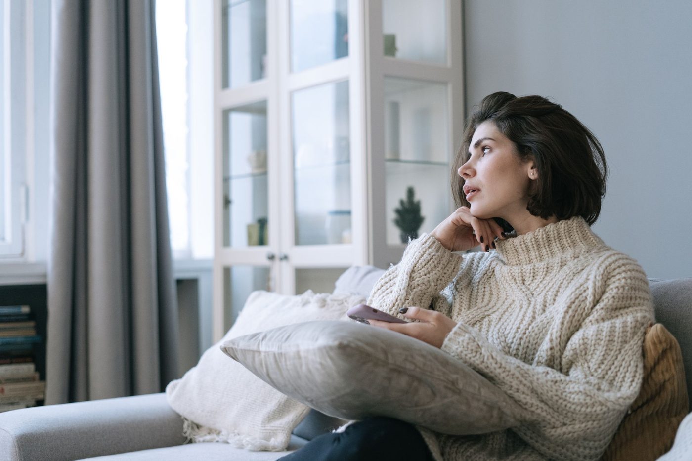 A woman in a cream knitted jumper sits on a sofa with her chin on her hand, looking out of a window with a phone in her hand