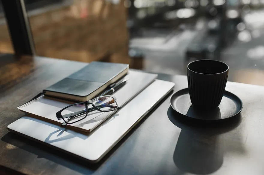 A closed laptop, notebooks, a pen and a pair of glasses beside a dark cup on a table by a sunlit window