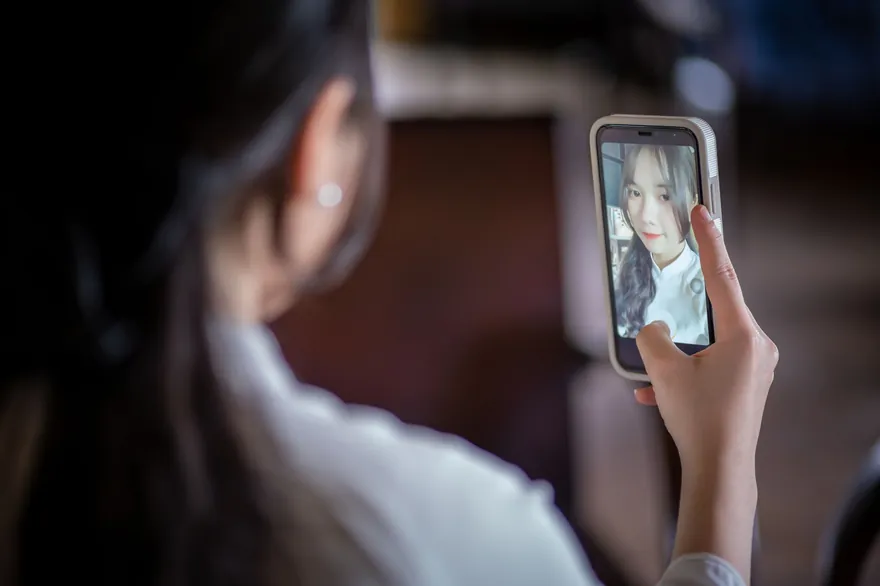 A woman seen from behind holds a phone up to take a selfie, her face on the screen