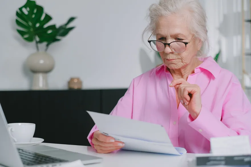 An older woman in glasses and a pink shirt reading a printed document at a desk, a cup and saucer beside her