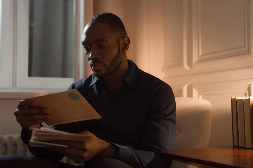 A man in a dark shirt reading a letter from a brown envelope in a softly lit room
