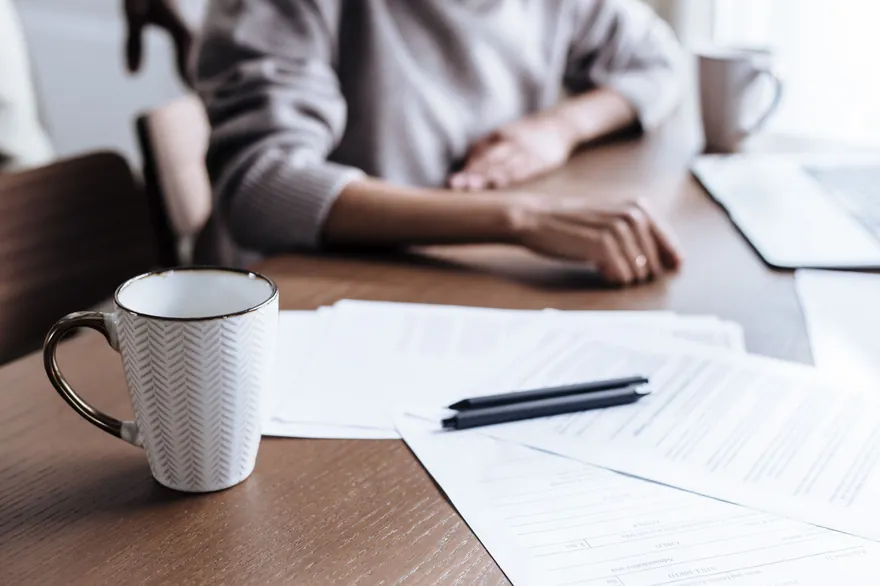 A mug, two pens and printed papers on a dark wooden table, with a person&rsquo;s arms resting on it