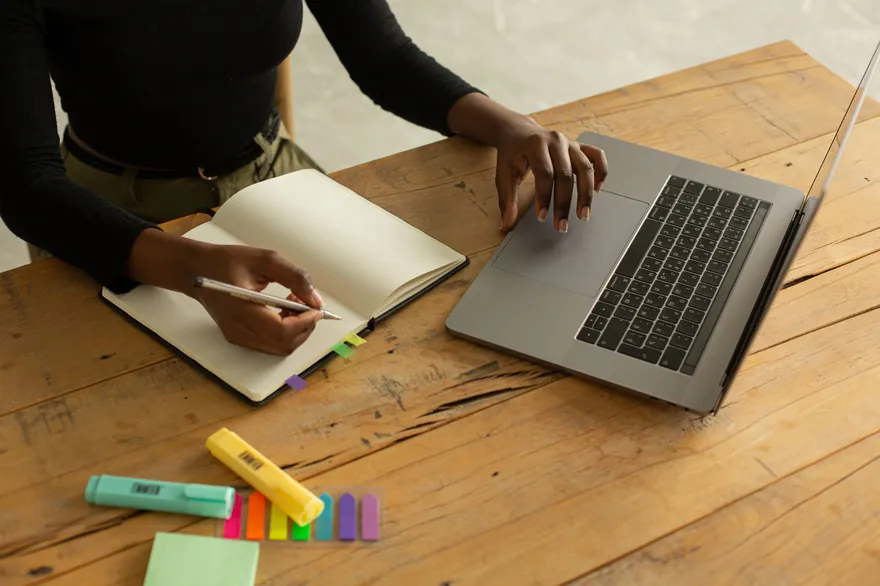 A person’s hands rest on an open notebook beside a laptop and highlighters on a wooden table