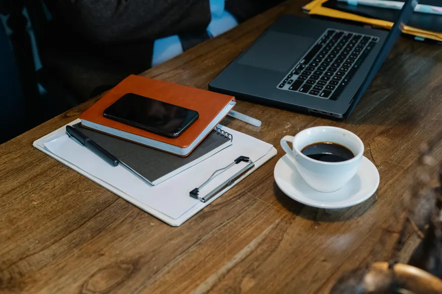 An open laptop, a notebook and a phone on a wooden desk, seen from above, with a cup of coffee