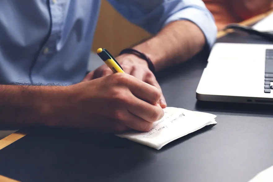A man&rsquo;s hand writes notes on a small pad beside a laptop on a dark desk