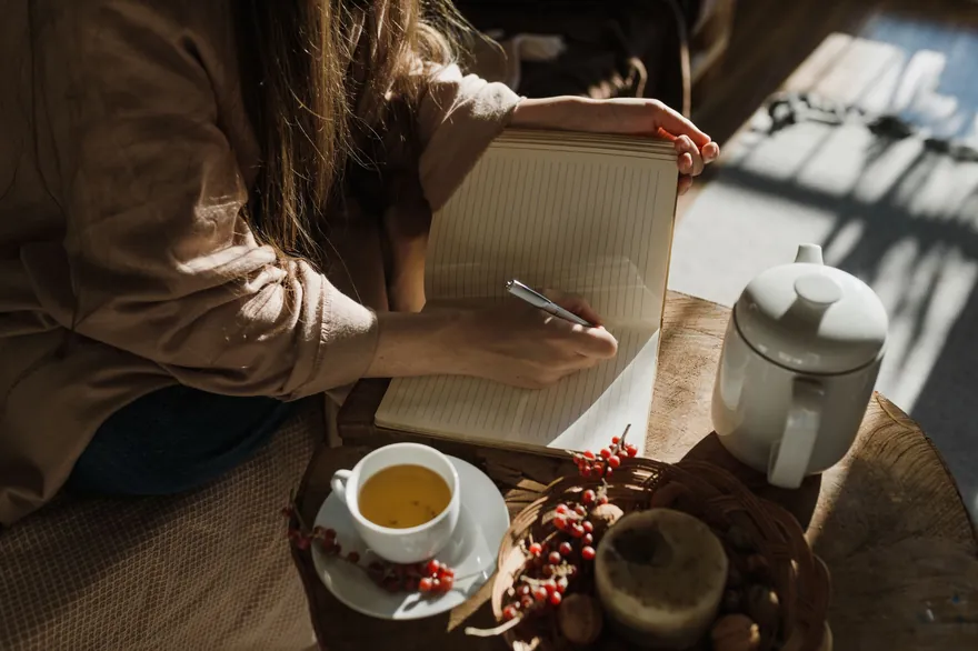 A woman writing in a lined notebook in low autumn sunlight, a cup of tea, a white teapot and a bowl of red berries on the table beside her