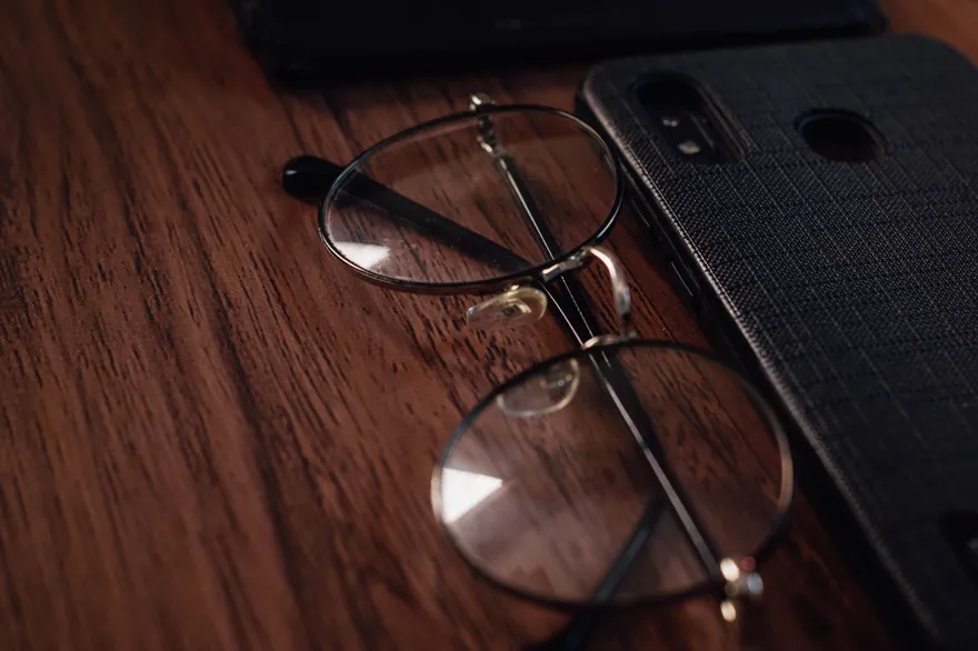 A phone in a fabric case lies on a wooden table beside a pair of round glasses