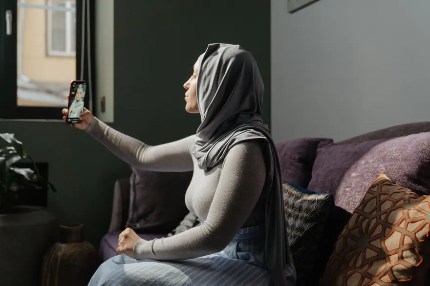 A woman in a grey headscarf takes a front-camera selfie on a sofa beside a window