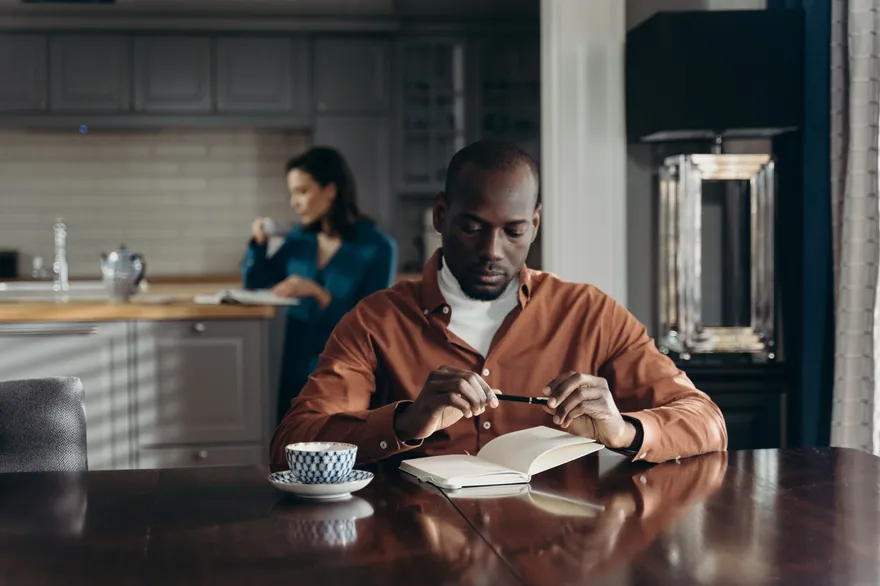 A man at a kitchen table holds a pen over an open notebook, a cup of tea beside him