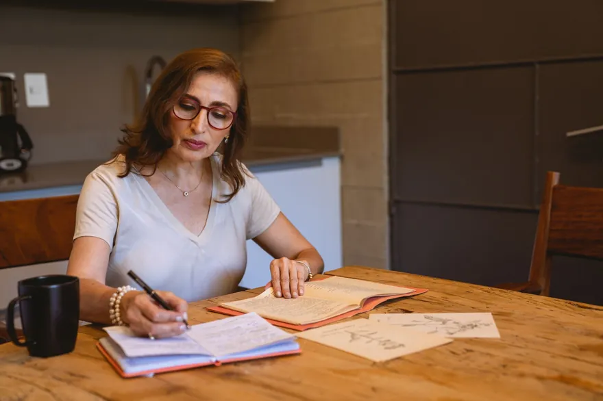 A woman in glasses writing notes into a notebook at a wooden kitchen table, a dark mug beside her