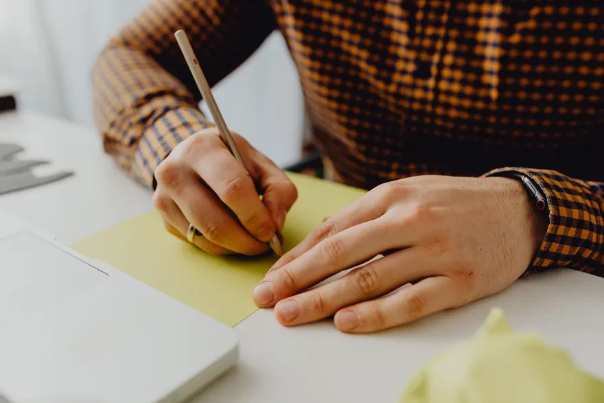 A hand writing with a pencil on a sheet of pale green paper at a white desk