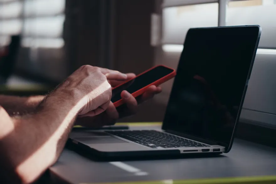 A hand holds a phone with a red case in front of an open laptop on a dark desk
