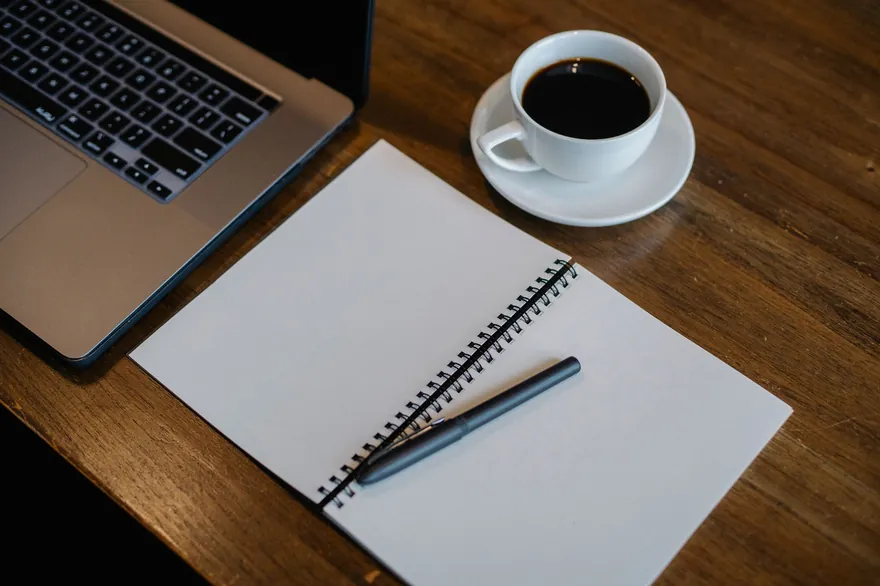An empty notebook page with a pen beside a laptop and a cup of black coffee on a wooden table