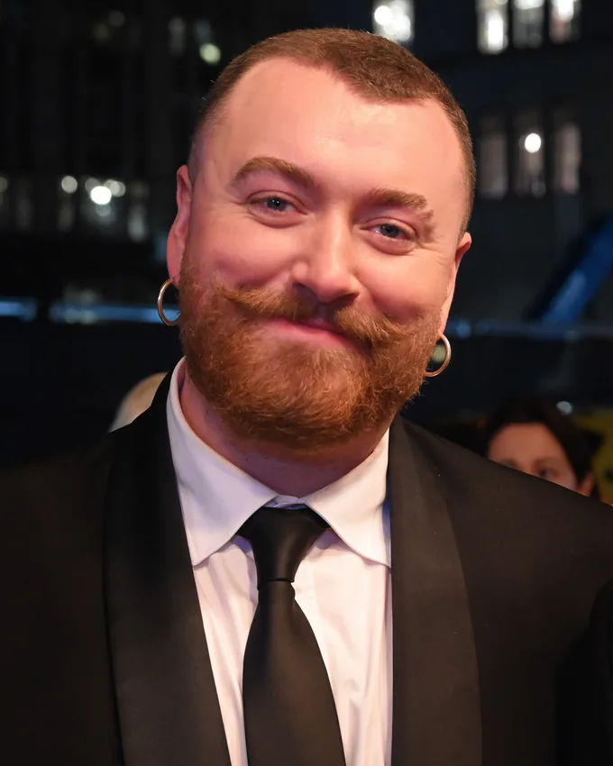 Sam Smith in a black suit and tie on a red carpet at night in 2025