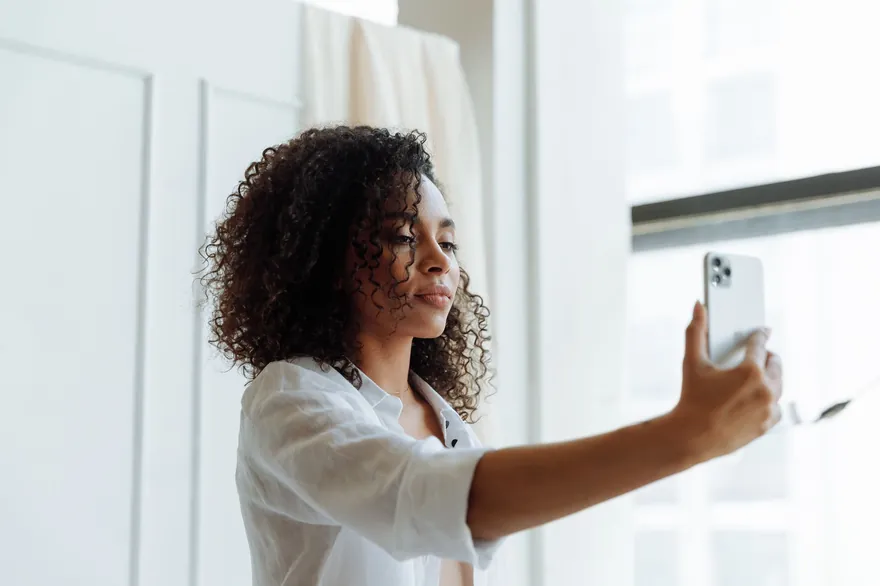 A woman with curly hair by a bright window holding a phone at arm’s length to film a selfie, the back of the phone towards the camera