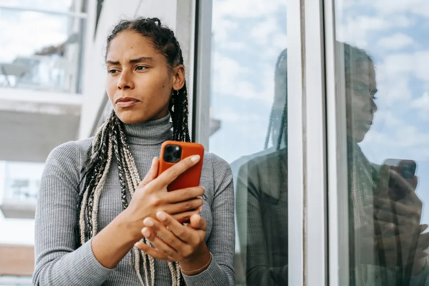A woman with braids in a grey roll-neck holds a phone and looks out of a window
