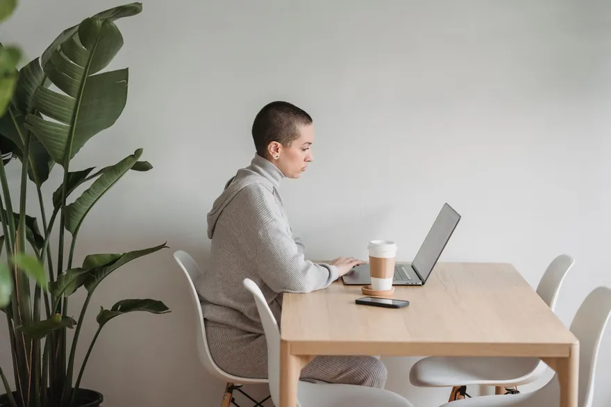 A person with short hair sits at a table in a bright living room, working on a laptop beside a plant