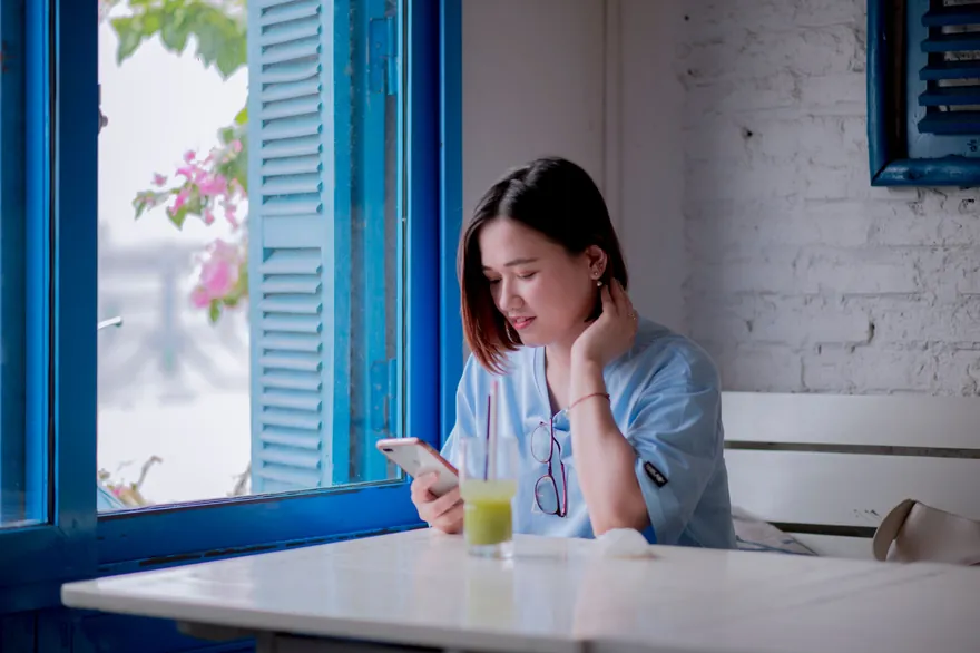 A young woman in a light blue shirt looks at her phone at a white table by a blue-framed window, a glass of juice beside her