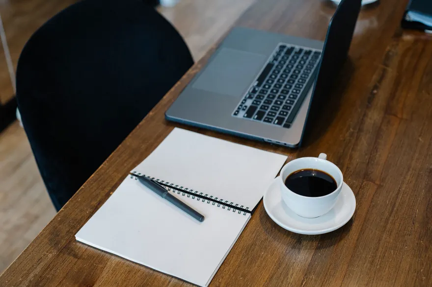 An open laptop and a spiral notebook with a pen and a cup of black coffee on a wooden table