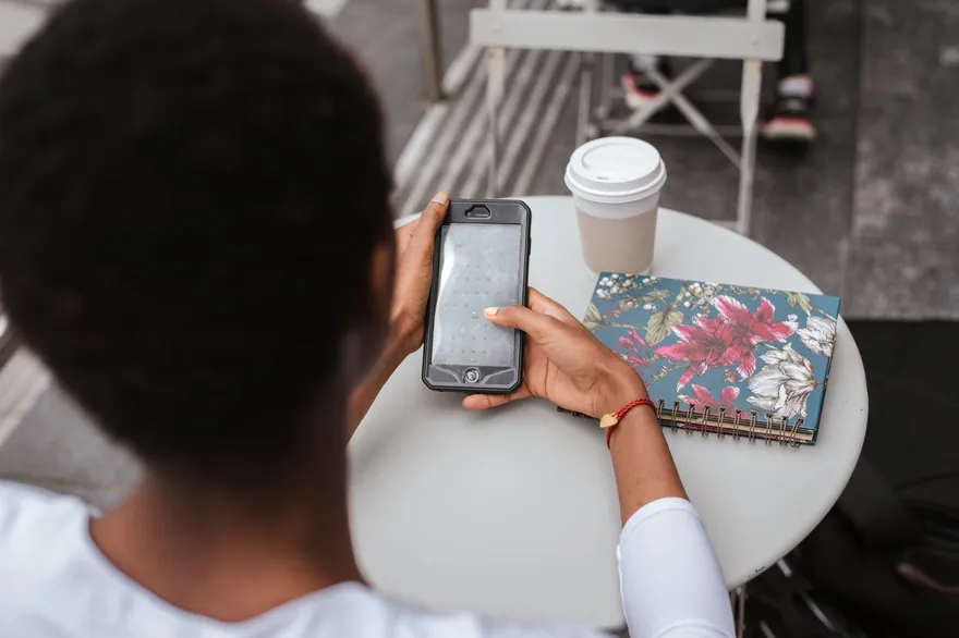 A person seen from behind holds a phone at a cafe table beside a notebook and a cup of coffee
