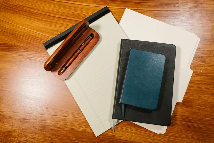 A notebook, a pad and folders with pens on a wooden desk, seen from above